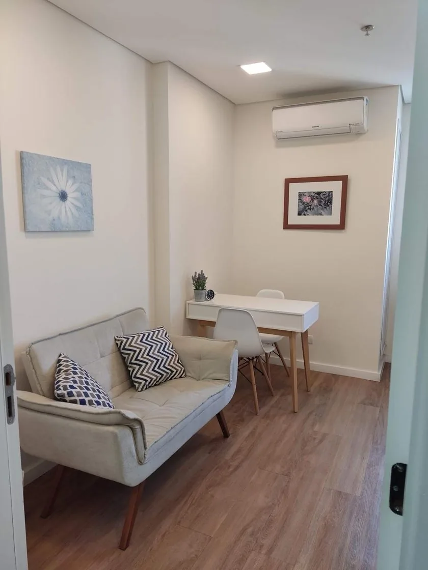 Consultório de Jaqueline Sousa em São Bernardo do Campo: sofá claro com almofadas, mesa branca com duas cadeiras, quadros na parede e piso de madeira
